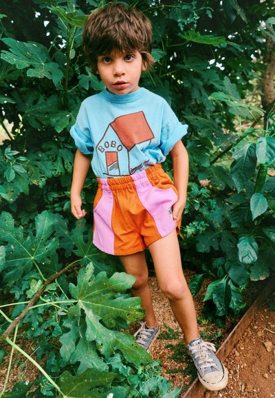 Jeune enfant aux cheveux bruns portant un t-shirt bleu et un short orange-rose, debout parmi des plantes vertes en plein air.