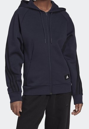 Osoba ubrana w granatową bluzę z kapturem zapinaną na zamek, z czarnymi paskami na rękawach i małą naszywką z logo Adidas na przedniej kieszeni.