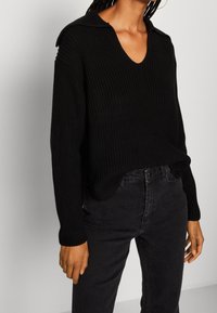 Maglione nero a maglia con scollo a V e design a spalle scese, tessuto testurizzato, abbinato a jeans neri, creando un look monocromatico.