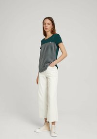 Top verde scuro a maniche corte con strisce bianche orizzontali e un piccolo logo, abbinato a pantaloni bianchi a gamba larga e sneakers beige.