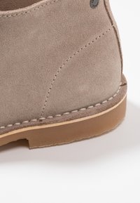 Gros plan sur le talon d'une chaussure en daim beige avec des coutures apparentes et une semelle en caoutchouc épaisse sur fond blanc.
