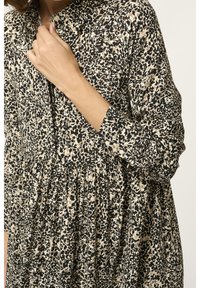 Femme portant une robe à manches longues, ample, avec un imprimé léopard abstrait noir, beige et blanc, main touchant le col près des boutons.