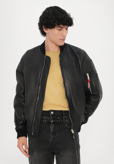 Black leather bomber jacket with ribbed cuffs and collar, featuring a zippered pocket on the sleeve and smooth finish. Worn over a yellow shirt.