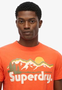 Orange bomulds t-shirt med et grafisk motiv af bjerge, træer og en sol, med ordet "Superdry" i fed, hvide bogstaver.