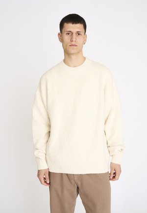 Junger Mann mit kurzen dunklen Haaren, der einen cremefarbenen, strukturierten Pullover und hellbraune Hosen vor einem schlichten weißen Hintergrund trägt.