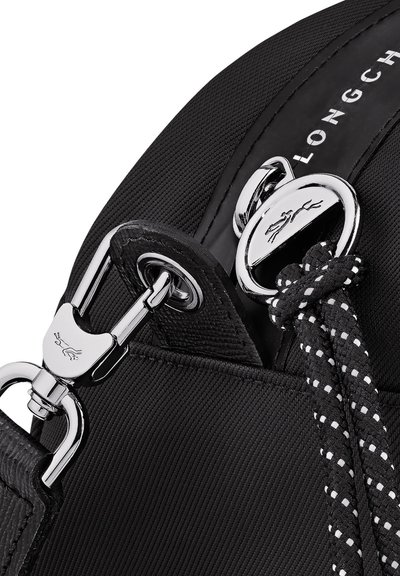 Nahaufnahme eines schwarzen Stofftaschenriemens mit silberner Metallklammer und Logo, der mit einem schwarz-weißen geflochtenen Kordel durch einen runden Metallring gebunden ist.