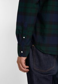 Camicia a maniche lunghe a quadri, con colori verde scuro e blu navy, dotata di polsini con bottoni e colletto a punta. Abbinata a jeans in denim scuro.