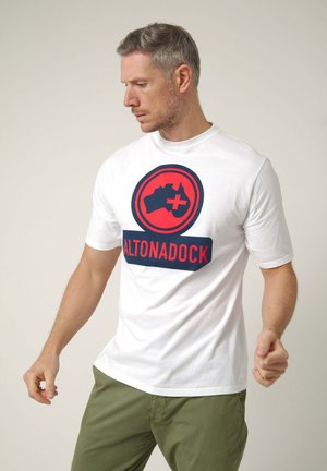 Hombre con camiseta blanca con el logo "ALTONADOCK" en rojo y azul marino y pantalones verde oliva, mirando hacia abajo y girando ligeramente el cuerpo.