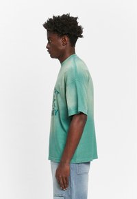 Mintgrøn oversized t-shirt med rund halsudskæring og korte ærmer, med et falmet gradientdesign og trykt grafik på fronten.
