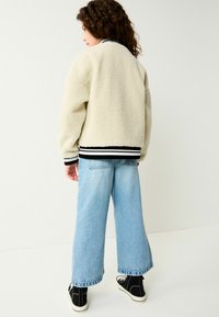 Kerman fleece bomber jacket, jossa on musta-valkoinen raitaresori ja hihansuut, yhdistettynä vaaleansinihin leveälahkeisiin farkkuihin ja mustiin korkeakorkoisiin lenkkitossuihin.