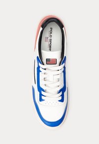 Zapatilla blanca con acentos azules y rojos, de cuero suave, con puntera redonda y suela de goma. Presenta la marca Polo Sport y un detalle de la bandera americana.