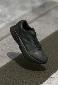 Chaussure de sport noire avec une tige texturée, présentant un design épuré, des lacets et une semelle rembourrée. Convient pour les activités de performance.