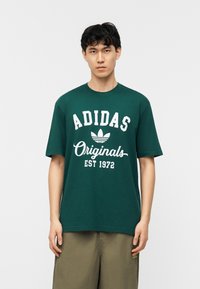 Νεαρός άνδρας που φοράει σκούρο πράσινο T-shirt Adidas Originals με λευκό λογότυπο και χακί σορτς, στέκεται μπροστά σε λευκό φόντο.