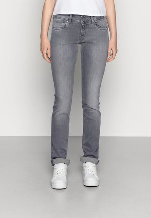 Person trägt graue Slim-Fit-Jeans mit aufgerollten Bündchen und weiße Sneakers, steht vor einem schlichten hellen Hintergrund.
