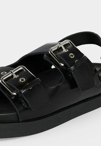 Μαύρο σανδάλι με δύο ασημένιες αγκράφες και χοντρή σόλα, εμφανιζόμενο σε κοντινή γωνία σε λευκό φόντο.