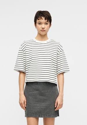 Νεαρό άτομο με κοντά σκούρα μαλλιά που φοράει μια λευκή και μαύρη ριγέ φαρδιά μπλούζα και μια σκουρόχρωμη γκρι τζιν φούστα.