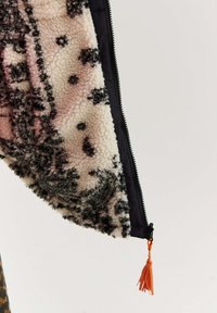 Nahaufnahme eines kuscheligen, gemusterten Jackenärmel mit einem schwarzen Reißverschluss und einem orangefarbenen Ziehvorgang vor einem schlichten Hintergrund.