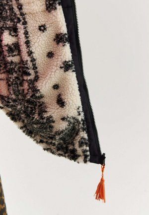 Detailní záběr na rukáv huňatého vzorovaného sáčka s černým zipem a oranžovým táhlejším páskem na pozadí.