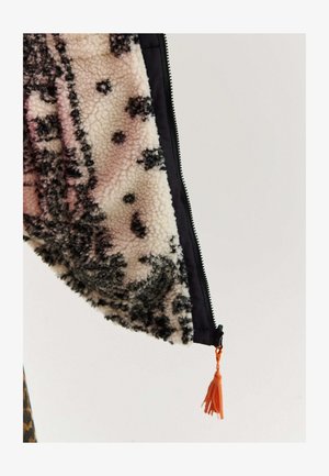 Detailný záber na rukáv huňatej vzorovanej bundy s čiernym zipsom a oranžovým strapcom na ťahanie na jednofarebnom pozadí.