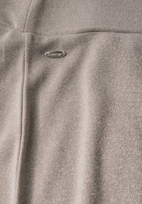 Beige tyg med en subtil lyster, som har en metallisk logotypaccent. Texturen är mjuk och vävd, vilket framhäver en slät och jämn yta.