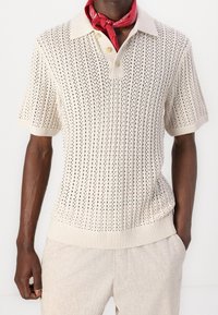 Polo de punto en crema con un patrón texturizado y transpirable. Presenta un cuello clásico, una tapeta de tres botones y puños y dobladillo acanalados.