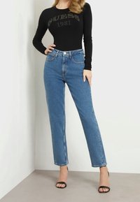 Bodysuit negro de manga larga con el logo "GUESS 1981", combinado con jeans azul de cintura alta y sandalias negras de tacón sobre un fondo neutro.