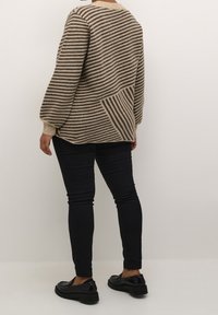 Pull beige et marron à rayures avec une coupe ample, un col rond et des manches texturées, associé à un jean skinny noir et des chaussures noires.