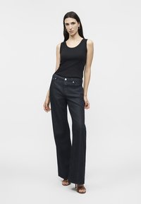 Femme portant un haut noir sans manches, un jean large sombre et des escarpins marron à bout ouvert, debout devant un fond clair uni.