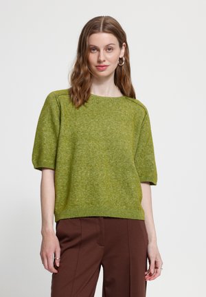 Femme aux cheveux bruns ondulés portant un pull en maille vert à manches courtes et un pantalon marron, debout devant un fond clair uni.