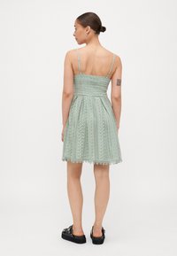 Világoszöld csipke ruha állítható pántokkal, testhez simuló felső résszel, bővülő szoknyával, mintás textúrával és fodros szegéllyel. Fekete cipővel viselve.