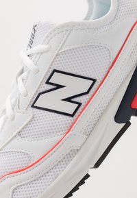 Bílý sportovní teniska s horní částí z síťoviny, černým logem a červenými doplňky. Má ploché tkaničky a texturovanou gumovou podrážku pro lepší přilnavost.