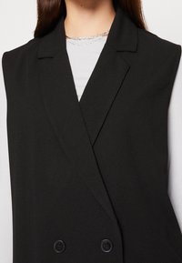 Svart ärmlös blazer med dubbelknäppt design, skarpa slag och mjukt tyg. Den har två stora svarta knappar för stängning.