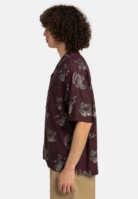 Kurzärmliges, kastanienbraunes Shirt mit lockerer Passform. Besitzt ein Frosch- und Skorpionmuster, leichtes Material und eine einzelne Brusttasche.