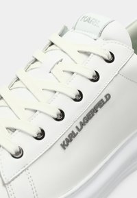 Balti odiniai sportbačiai su tekstūruota paviršiumi; turi plokščias baltas raišteles, sidabrinės spalvos logotipą šone ir apvalų pirštą.