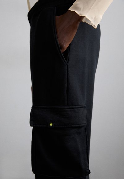 Pantaloni cargo neri con tasca laterale, realizzati in un tessuto texturizzato e con un risvolto. Dettaglio del pulsante giallo ben visibile sulla tasca.