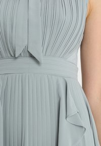 Harmaa mekko, jossa on pleated-tekstuuri, sidottu kaulustyyli ja liehuva hame, jossa on frillakoristelu helmassa.