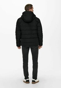 Black puffer jacket with a hood, quilted design, and elastic cuffs. Worn with grey jeans and white sneakers with black accents.
