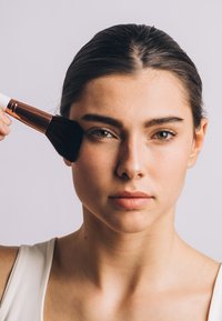Makeup børste med hvit håndtak og rosagull ferrule, som påfører blush på en hudfarget ansikt, med jevn hud og naturlig sminke.