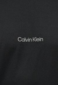 Μαύρο υφασμάτινο με υφή, με το λογότυπο «Calvin Klein» σε λευκό χρώμα τυπωμένο στο κέντρο του υλικού.