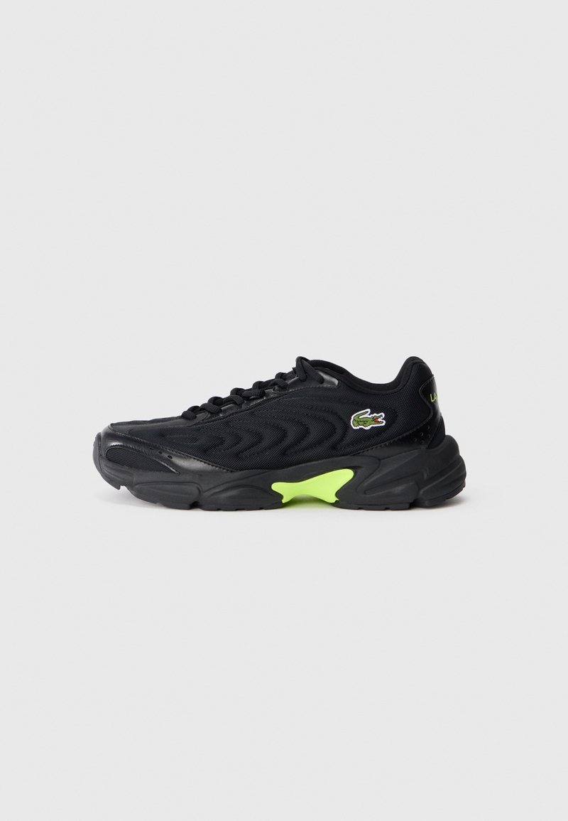 Baskets noires avec un design en mesh ondulé, fermeture à lacets, détail de semelle vert néon et un petit logo crocodile vert sur le côté.