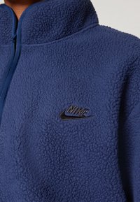 Nike fleecejacka i marinblått, med hög krage, dragkedjeslutning och svart broderad logga. Strukturerat, mjukt material utan mönster.
