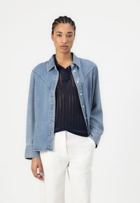 Giacca di jeans di colore azzurro chiaro, con colletto classico e chiusura a bottoni, sovrapposta a una blusa trasparente blu navy, abbinata a pantaloni bianchi.