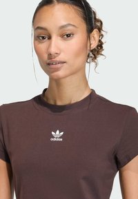 Hnedé bavlnené tričko s krátkymi rukávmi a okrúhlym výstrihom, s bielym logom Adidas na hrudi, hladkou textúrou a priliehavým dizajnom.