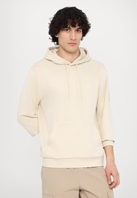 Felpa beige con cappuccio, tasca frontale, vestibilità ampia, polsini a coste e una sottile etichetta con logo sulla manica. Tessuto morbido e testurizzato.