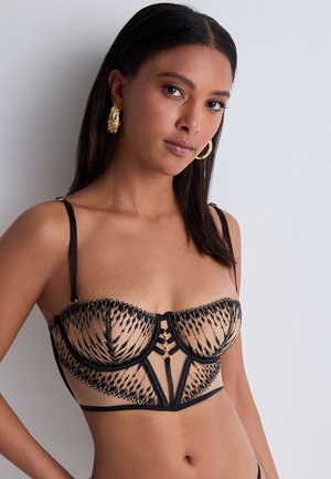 Soutien-gorge en dentelle noire avec une base beige transparente, présentant des motifs rayés complexes, des accents en métal doré et des bretelles réglables.