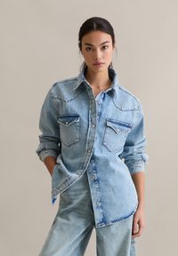 Lichtblauw denim overhemd met knoopsluiting aan de voorkant, twee borstzakken en opgerolde mouwen. Voorzien van een klassieke kraag en subtiele stiksel details.
