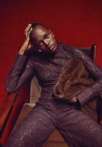 Mono de encaje púrpura con mangas largas y cuello alto, sostenido por un accesorio de piel sintética marrón, sentado en una silla de madera contra un fondo rojo.