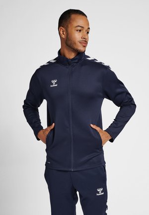 Marineblaue Sportjacke mit Reißverschluss und hohem Kragen, aus glattem Stoff gefertigt, mit weißen Zickzackstreifen auf den Schultern und einem Logo auf der Brust.