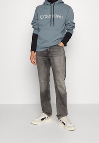 Grå bomullströja med vit "Calvin Klein" logotyp, i kombination med urtvättade grå jeans och vita sneakers med marinblå detaljer. Tröjan har en känguruficka.