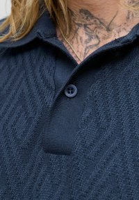 Tamsiai mėlynas polo marškinėliai, pagaminti iš tekstūrinio mezginio audinio su zigzago raštu, su dviem knopkėmis ir apykakle.